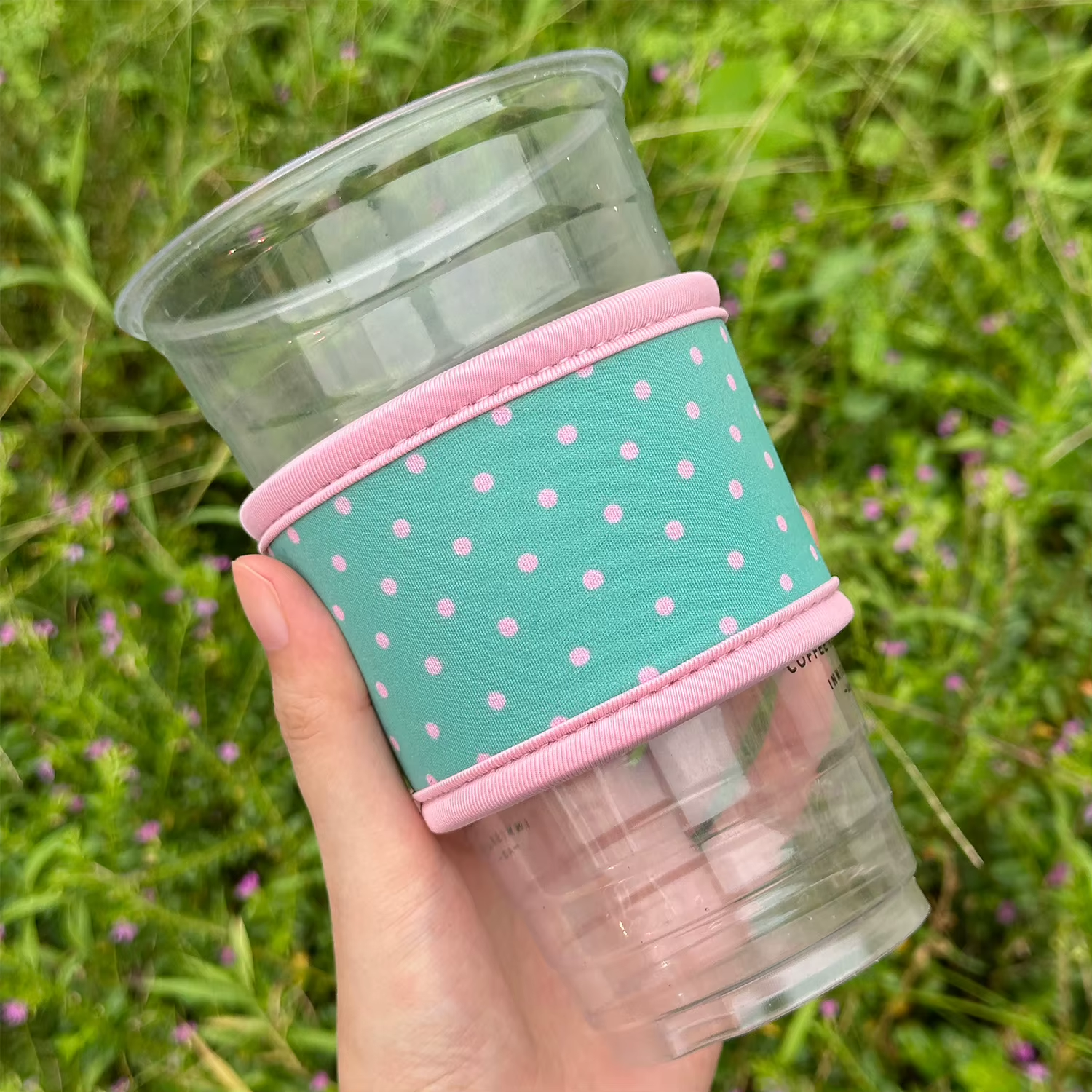 Hand supporting a clear cup in a shorter teal bottomless coffee sleeve with pink polka dots and pink upper and lower binding; cup base exposed.